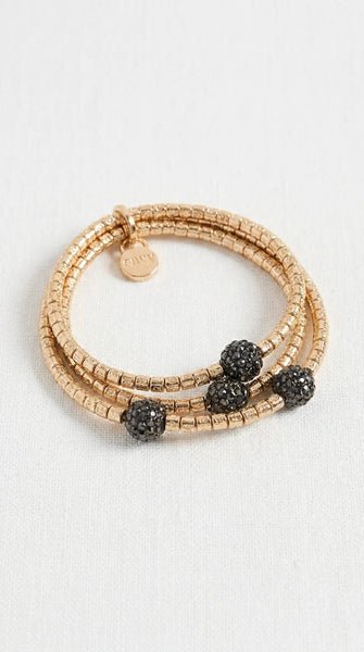 Envy gold beaded wrap bracelet with three textured black crystal ball charms, displayed on a light off-white fabric background.