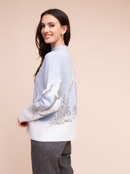 A woman wearing a light blue and white knit sweater with silver-grey abstract patterns on the sleeves and back, paired with gray trousers, set against a solid beige background.