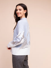A woman wearing a light blue and white knit sweater with silver-grey abstract patterns on the sleeves and back, paired with gray trousers, set against a solid beige background.
