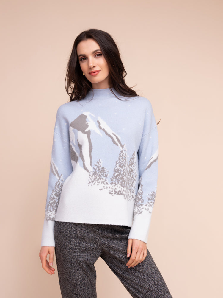 A woman modeling a light blue and white knit sweater with a mountain and tree pattern, paired with dark grey pants, against a solid beige background.