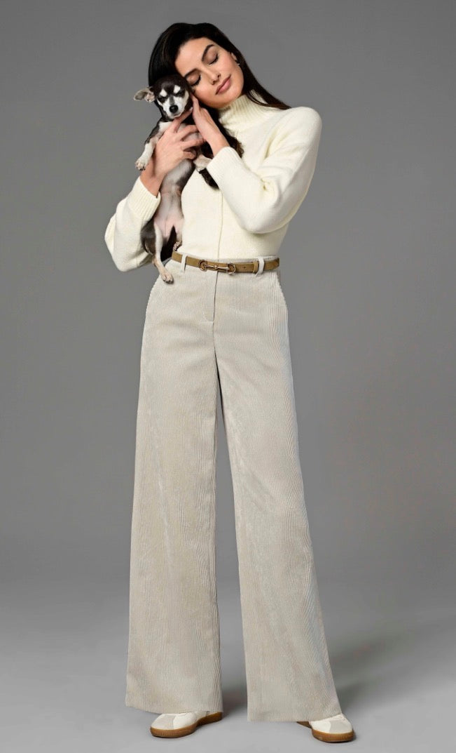 Model wearing Robell beige flared long length trousers with a tan belt. With a cream roll neck sweater and white sneakers. Cuddling a small dog on a grey studio background.