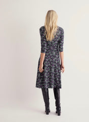 A woman viewed from behind wearing Seasalt  knee-length floral dress with three-quarter sleeves and black knee-high boots, against a plain light beige studio background.