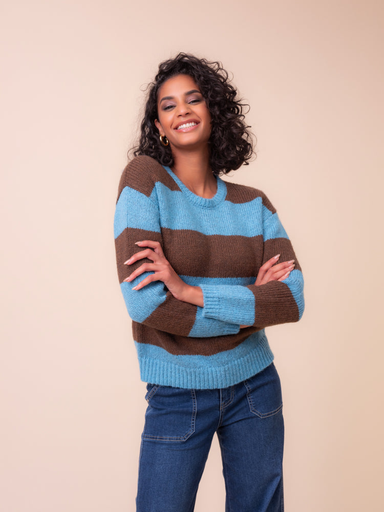 A woman with curly black hair wearing Alison Sheri chunky knit brown and  blue striped sweater and dark blue jeans. Standing against a solid beige background with her arms crossed.