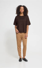 A woman modelling an Ichi dark brown short-sleeve top, with tan trousers against a plain light grey studio background.