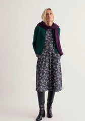 A woman modelling a Seasalt dark floral midi dress with a green cardigan, a deep purple scarf, and black knee-high boots, photographed against a plain light grey studio background.