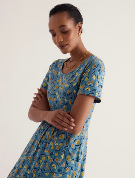 A woman wearing Seasalt short sleeve blue dress with a yellow and green floral pattern, photographed against a plain light background.