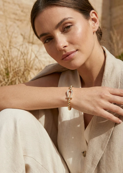 Model wearing a beige outfit with an Envy gold beaded bracelet with gold pendant with diamanté detail. In a garden setting.