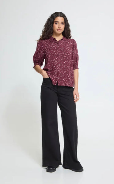 Model wearing Ichi floral short sleeve shirt in burgundy with black trousers and black shoes on a light grey background. 