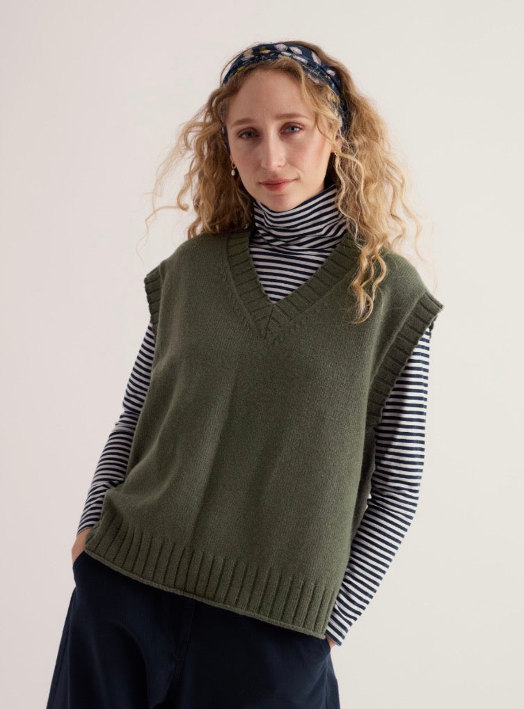 A woman wearing Seasalt olive green knit vest over a black and white striped turtleneck, paired with dark navy pants and a navy patterned headband, photographed against a plain light background.