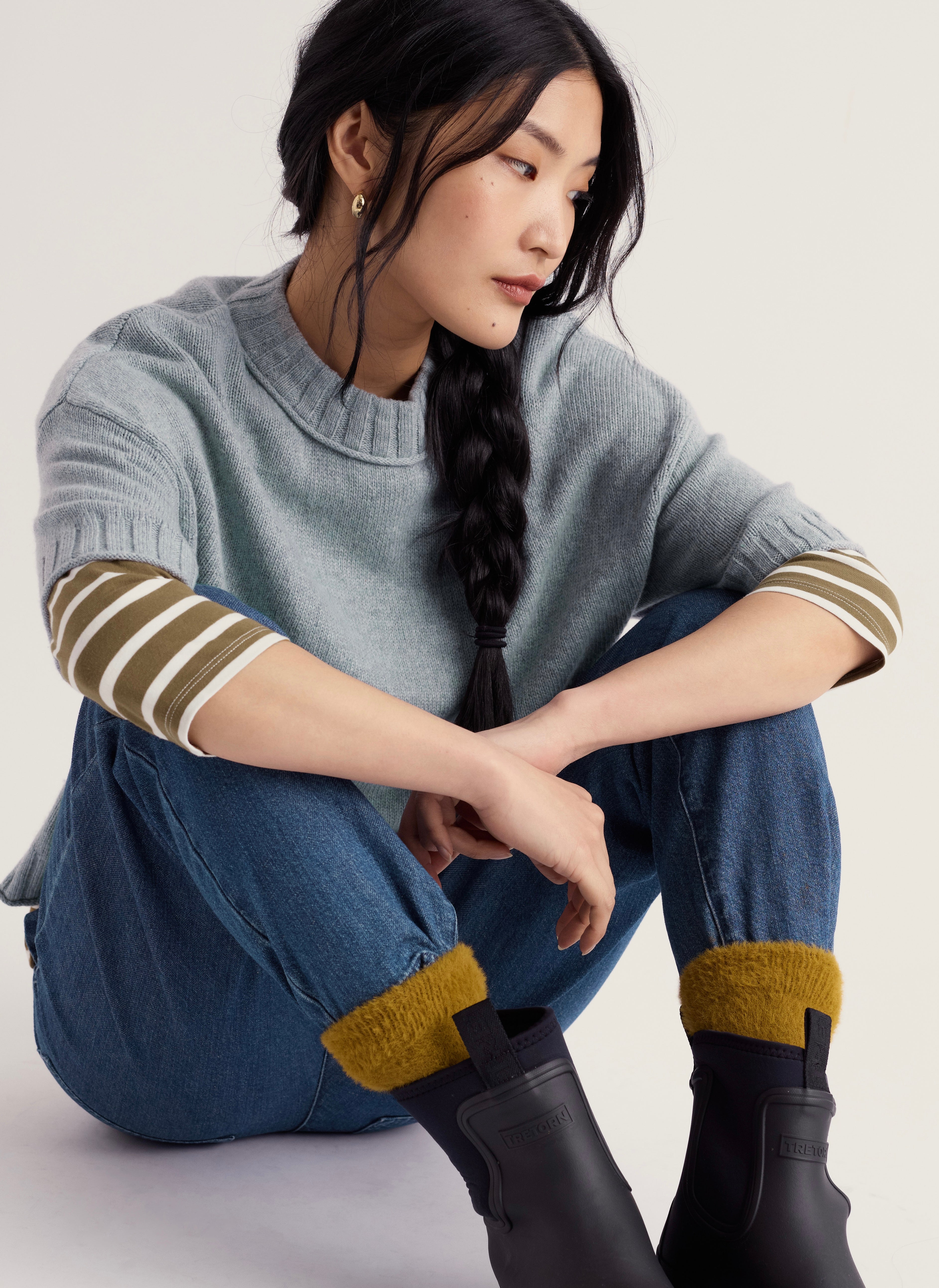 Model sat on white background floor wearing Seasalt light blue crew neck short sleeve jumper. With striped olive green top with blue jeans, mustard socks and black boots.