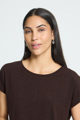 A woman with long dark hair wearing a dark brown short-sleeve Ichi t-shirt, shown from the chest up against a light gray studio background.