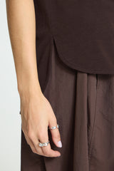 Close-up of a person's hand wearing two silver rings, dressed in a dark brown top and matching wide-leg skirt or trousers against a light gray background.