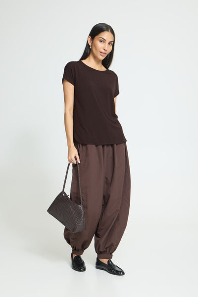 A woman modeling a dark brown short-sleeve top and matching loose-fitting brown pants, holding a dark brown textured handbag against a plain light gray studio background.