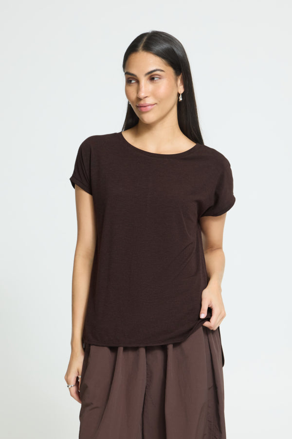 A woman modeling a dark brown short-sleeve Icho T-shirt and matching brown pants against a light gray studio background.