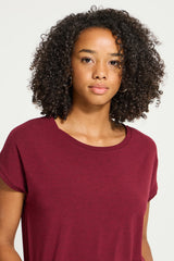 A woman with curly dark hair wearing a short-sleeve crewneck t-shirt in heathered burgundy, photographed against a clean off-white background.