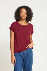A woman with curly dark hair wearing a short-sleeve crewneck Ichi t-shirt in burgundy and blue jeans, standing against a plain light gray studio background.