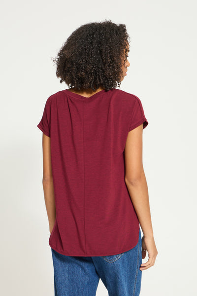 A woman viewed from the back wearing a short-sleeve maroon t-shirt and blue jeans against a plain white background.