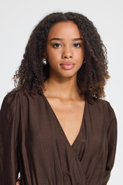 A woman with curly dark hair wearing a dark brown wrap-style Ichi Dress, photographed against a plain light gray studio background.