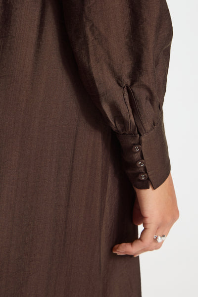 Close-up of a dark brown long-sleeve dress or tunic with three decorative buttons on the cuff, worn by a model whose hand and part of the body are visible against a plain light background.
