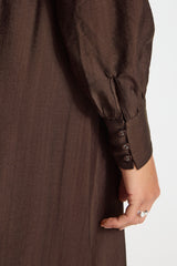 Close-up of a dark brown long-sleeve dress or tunic with three decorative buttons on the cuff, worn by a model whose hand and part of the body are visible against a plain light background.
