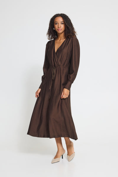 A woman modeling a dark brown Ichi midi wrap dress with long sleeves, a V-neckline, and a drawstring waist, paired with beige pointed-toe heels against a plain white studio background.