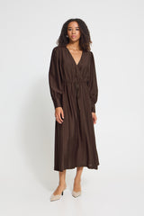 A woman modeling a dark brown long-sleeve midi dress with a V-neckline and drawstring waist, paired with beige pointed-toe heels, against a plain light gray studio background.