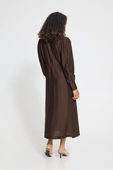 A woman viewed from the back wearing a dark brown long-sleeve midi dress with a gathered waist, paired with beige high-heeled sandals, against a plain white studio background.