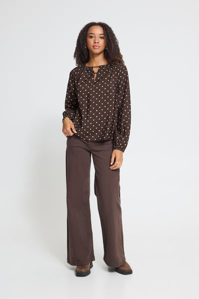 A woman modeling a dark Ichi brown long-sleeve blouse with a small white polka dot pattern and matching wide-leg brown trousers, standing against a light gray studio background.