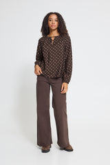 A woman modeling a dark Ichi brown long-sleeve blouse with a small white polka dot pattern and matching wide-leg brown trousers, standing against a light gray studio background.