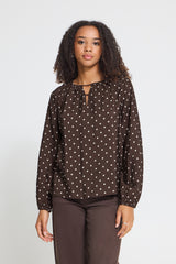 A woman modeling a dark brown long-sleeve blouse with a white polka dot pattern and a keyhole tie neckline, paired with dark brown pants, against a light gray studio background.