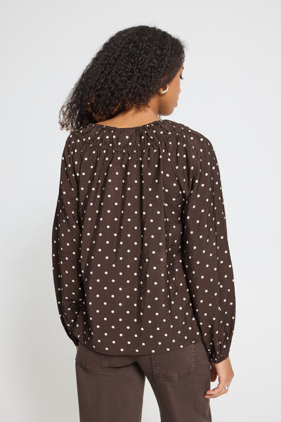 A woman shown from the back wearing a dark brown long-sleeve blouse with a white polka dot pattern and dark brown pants, set against a plain light gray studio background.