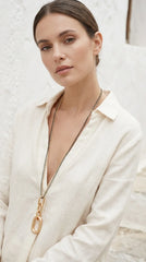 A woman wearing a cream-coloured linen shirt and an Envy long  gold chain necklace with a large gold ring pendant, set against a textured white wall.