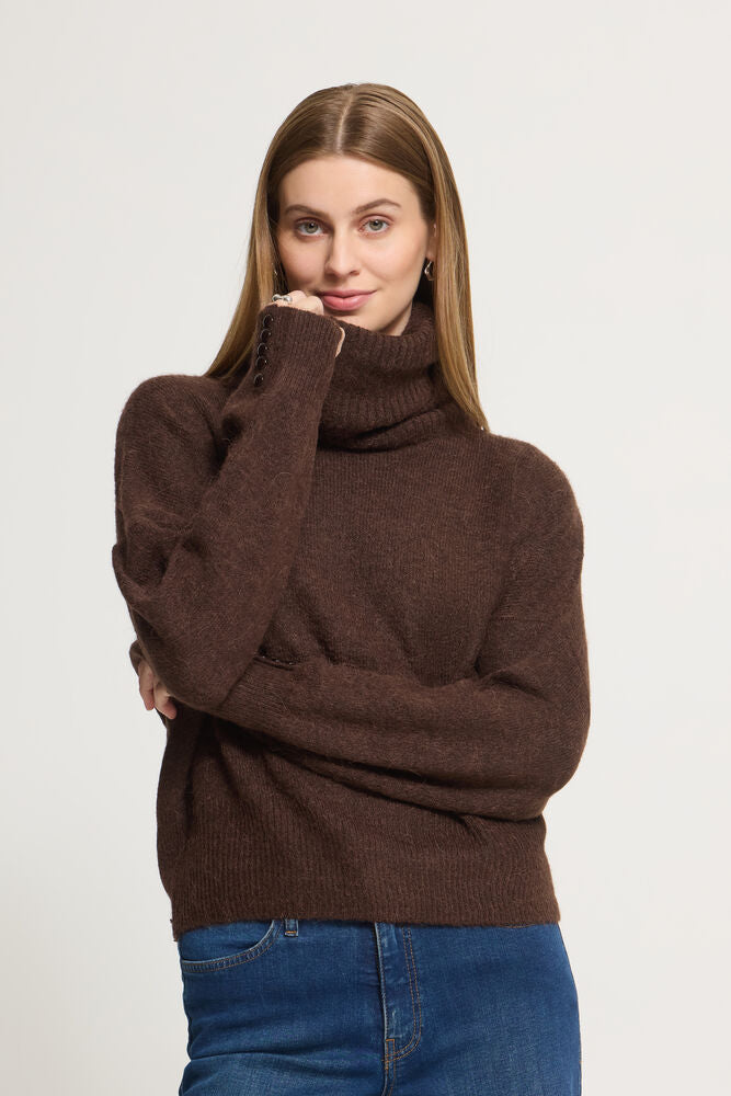 A woman wearing a dark brown Ichi  roll neck sweater and blue jeans, posing against a light grey  studio background.