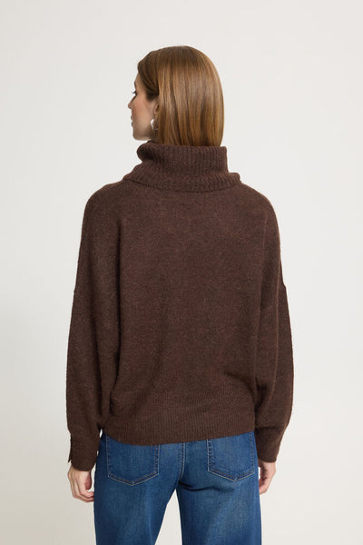 A woman viewed from the back wearing a Ichi dark brown ribbed turtleneck sweater and blue jeans, standing against a plain light grey studio background.