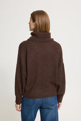A woman viewed from the back wearing a Ichi dark brown ribbed turtleneck sweater and blue jeans, standing against a plain light grey studio background.