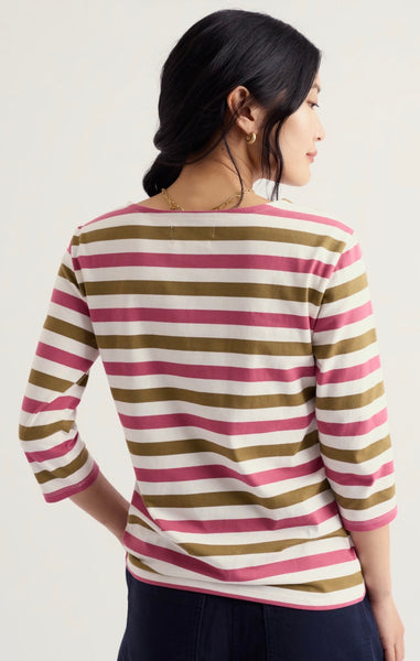 A woman shown from the back wearing a Seasalt 3/4 sleeve striped top with horizontal bands of pink, olive green, and cream, paired with dark navy pants, against a light off-white background.