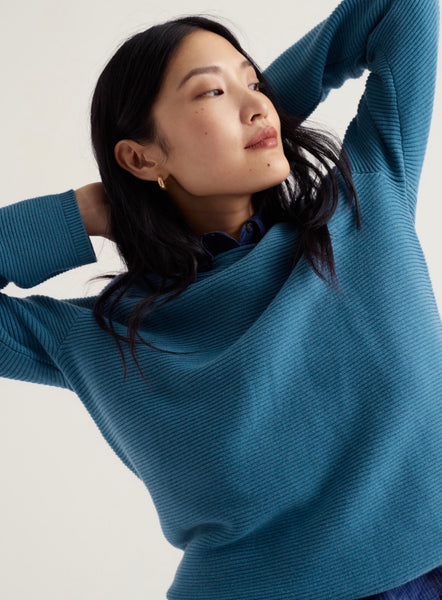 A woman wearing Seasalt ribbed knit blue sweater with a relaxed, draped neckline, posed with her arms raised against a plain light background.t 