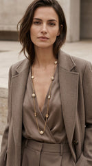 A woman wearing a taupe-brown double-breasted blazer over a matching satin top and trousers, accessorized with an Envy Jewellery long gold coloured chain necklace featuring round pearl-like beads.