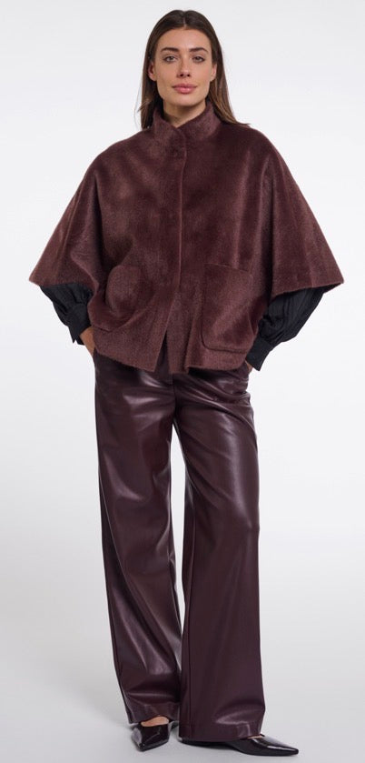 A model wearing Rino & Pelle dark brown textured cape with black sleeves and matching dark brown wide-leg pants, standing against a light grey studio background.