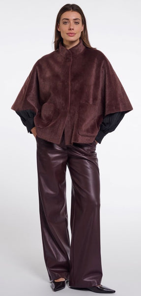 A model wearing Rino & Pelle dark brown textured cape with black sleeves and matching dark brown wide-leg pants, standing against a light grey studio background.