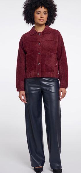 A model wearing a Rino & Pelle burgundy cropped jacket and shiny black wide-leg trousers, standing against a light grey studio background. 