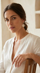 A woman with dark hair tied back wears a white linen shirt and textured Envy gold disc earrings, leaning on a wooden chair in a warm, softly lit interior.