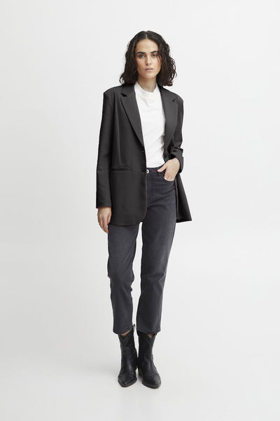 A woman modelling Ichi oversized black blazer over a white top, paired with dark grey pants and black ankle boots, standing against a light grey studio background.