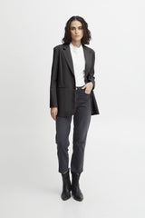 A woman modelling Ichi oversized black blazer over a white top, paired with dark grey pants and black ankle boots, standing against a light grey studio background.