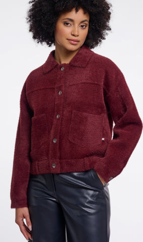 A model from the back wearing a Rino & Pelle, cropped maroon button-up jacket and shiny black leather pants, holding a brown leather shoulder bag against a plain white background.