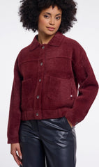 A woman wearing a maroon Rino & Pelle button up jacket with a collar and front pockets, paired with black faux leather pants, against a plain light grey background.