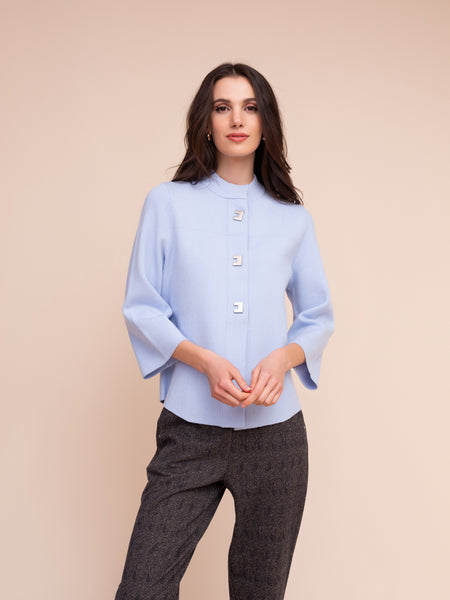 A woman modeling a light blue mock-neck blouse with three-quarter bell sleeves and decorative buttons down the front, paired with dark grey textured trousers, set against a solid beige background.