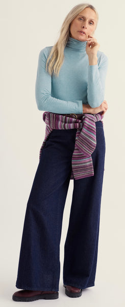 A woman modelling a Seasalt light blue  roll neck top, dark wide-leg denim pants, and a purple striped knit scarf tied around her waist, set against a plain off-white studio background.