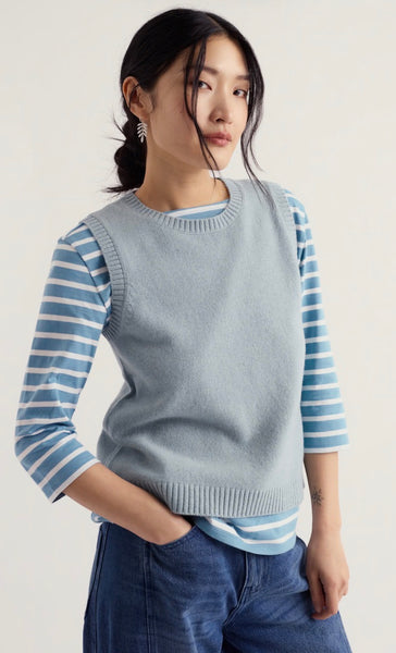 A woman modelling Seasalt vest top with a striped sleeved top underneath and wearing jeans with her hand in the front pocket in a studio background.