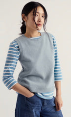 A woman modelling Seasalt vest top with a striped sleeved top underneath and wearing jeans with her hand in the front pocket in a studio background.
